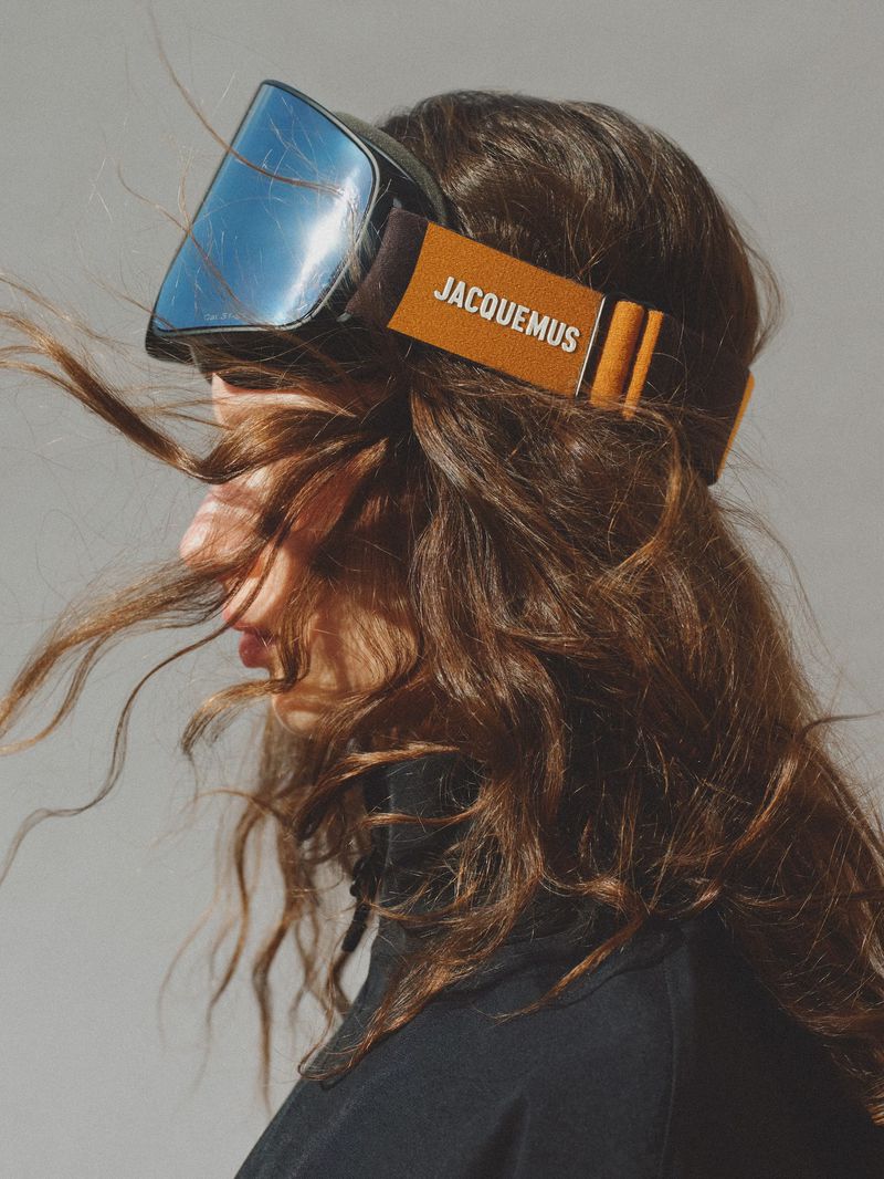 Profile portrait of a woman with curly hair and ski goggles with 'JACQUEMUS' written on the band, shot in a photo studio.