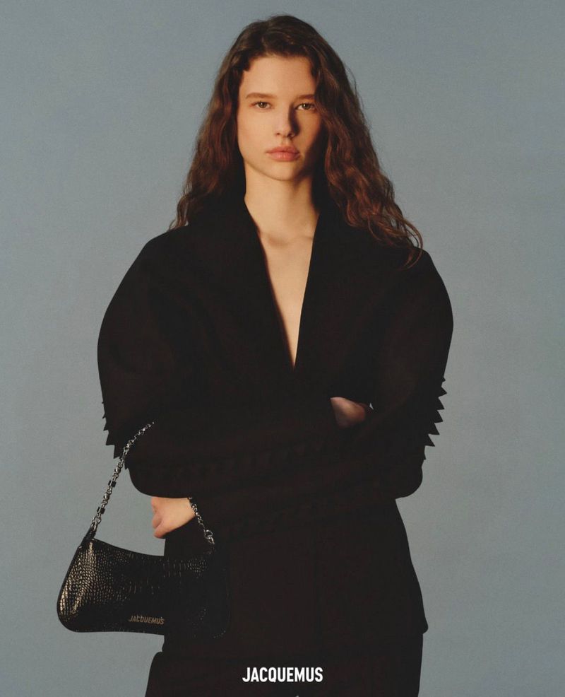 Model Sandra Murray posing in a sculpted black plunge blazer holding a black shoulder bag against a plain grey studio backdrop.