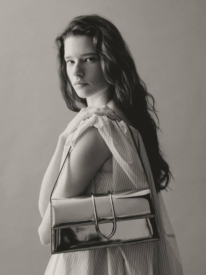 Monochrome fashion portrait of a woman with long dark hair, wearing a light dress and a shiny silver rectangular shoulder bag.