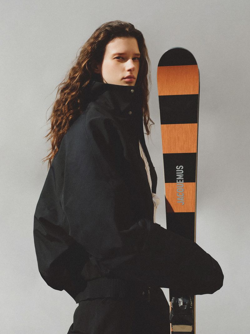 A fashion shot features a stylish woman with long brown hair in a black jacket, posing indoors next to a pair of black and brown skis.