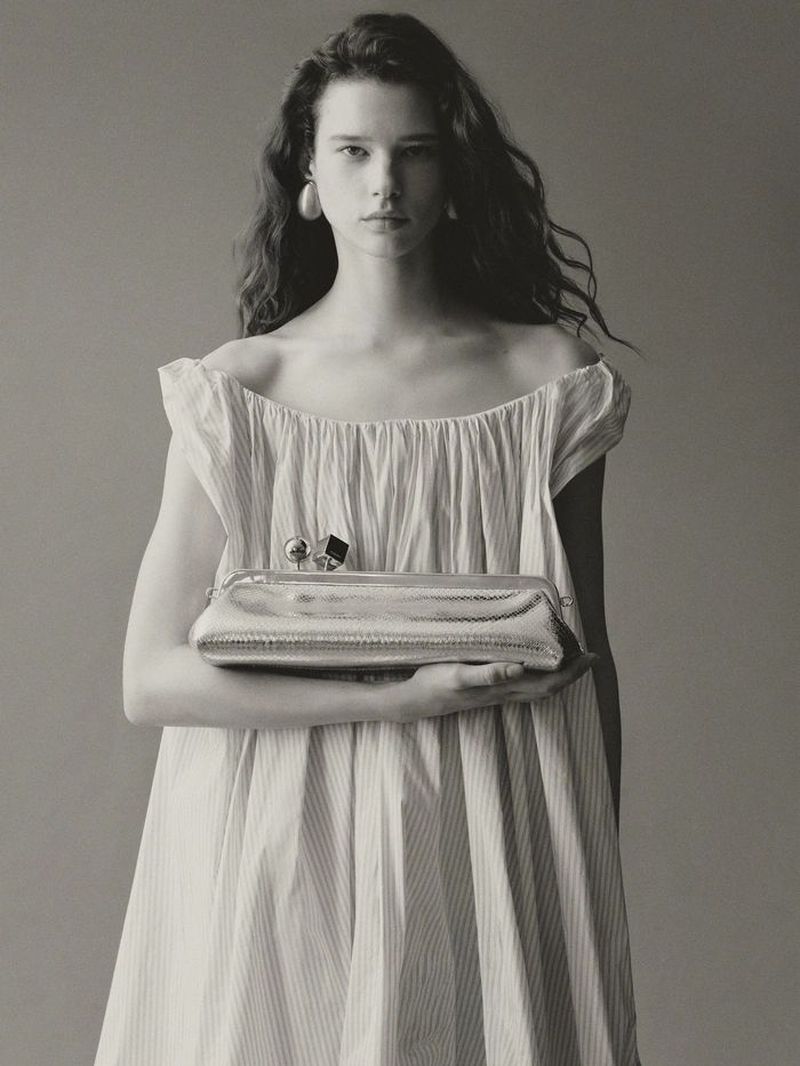 Studio shot of a woman holding a shiny silver clutch. She is wearing a white dress with off-the-shoulder sleeves and posing against gray backdrop.
