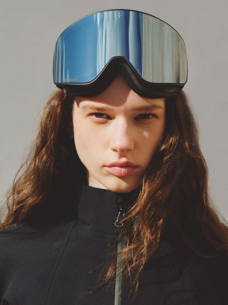 Portrait of a young woman wearing a black zip-up jacket and blue mirrored ski goggles against a neutral background.