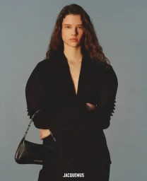 Model Sandra Murray posing in a sculpted black plunge blazer holding a black shoulder bag against a plain grey studio backdrop.