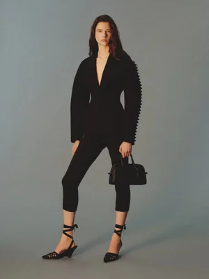 Model posing in a structured black blazer with pleated sleeves, cropped trousers, and ankle-tie heels against a grey seamless backdrop.