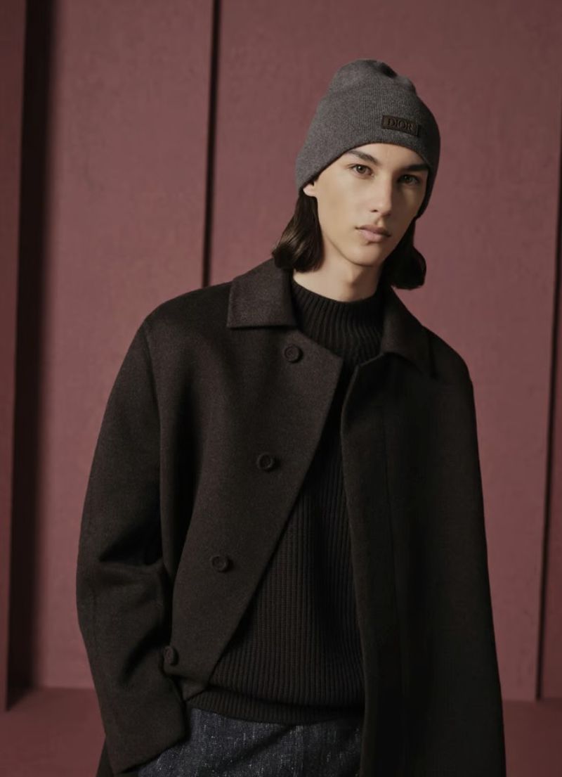 Studio shot of a male model wearing a beanie and dark overcoat against a dark pink studio backdrop, showcasing fashion and style.