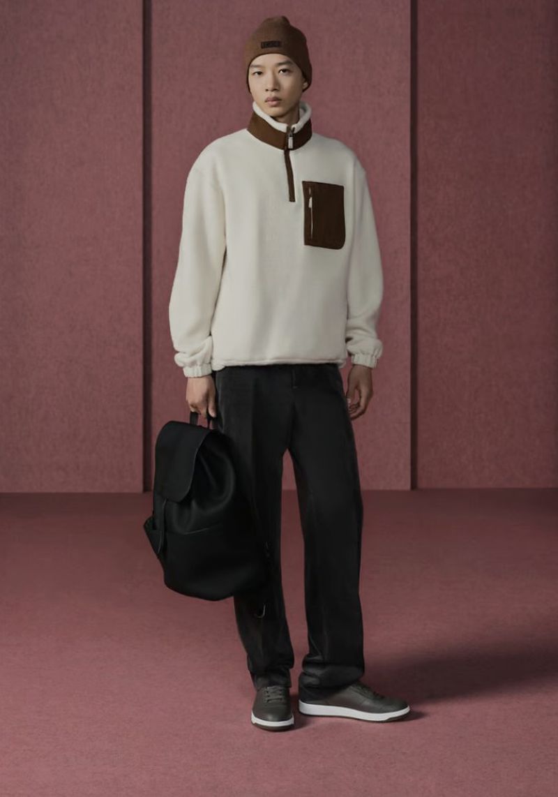 Full body shot of a young adult in a cream-colored sherpa pullover, dark pants and shoes, and a brown beanie, holding a black backpack.