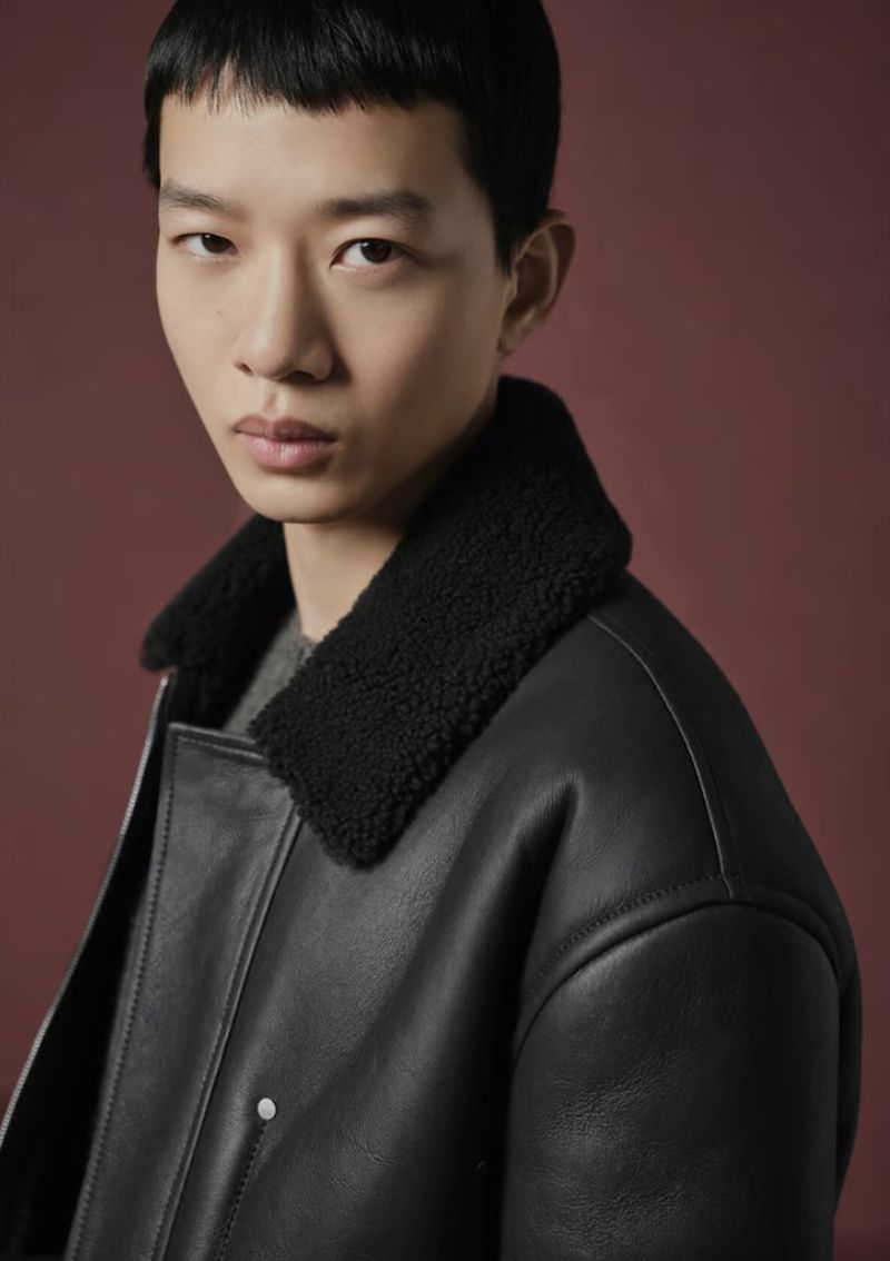Close-up of an Asian man in a stylish black leather jacket with a fur collar against a solid reddish-brown backdrop.