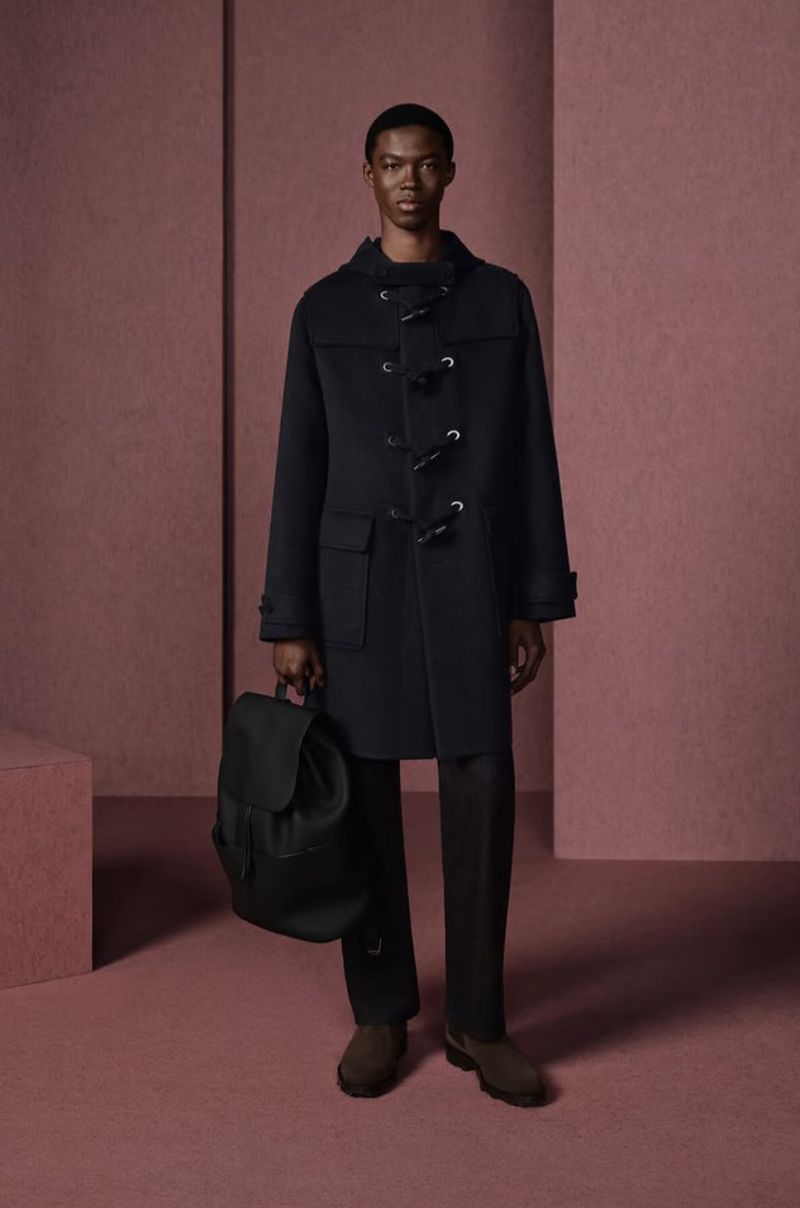 A solitary Black male model poses in a photo studio. He is wearing a toggle coat and holding a black backpack, with a monochromatic background.