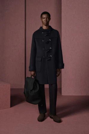 A solitary Black male model poses in a photo studio. He is wearing a toggle coat and holding a black backpack, with a monochromatic background.