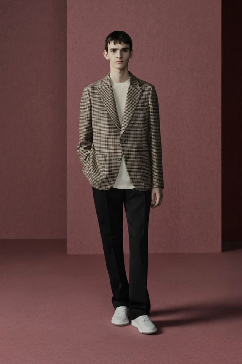 A full-length studio shot of a slender adult male wearing a patterned blazer, t-shirt, dress pants, and tennis shoes against a mauve backdrop.