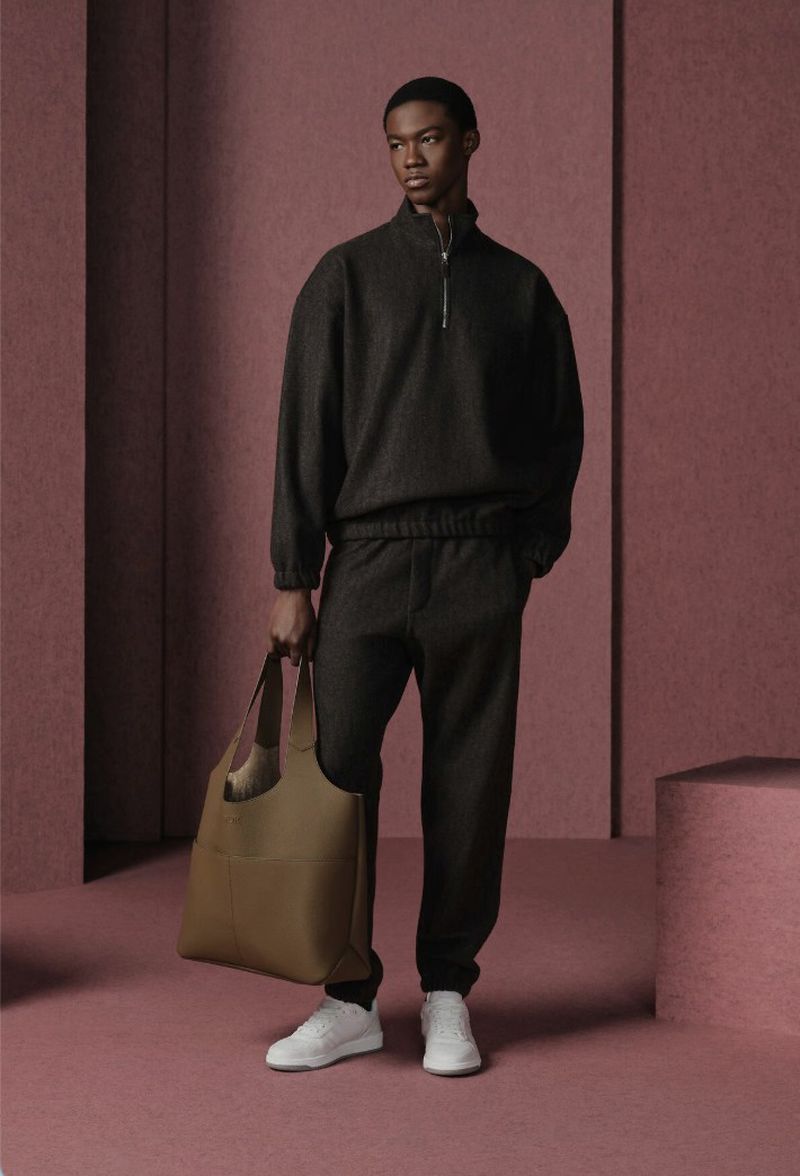 A dark-skinned man in a grey two-piece outfit holding a light-brown tote bag stands in front of a reddish-brown architectural background.