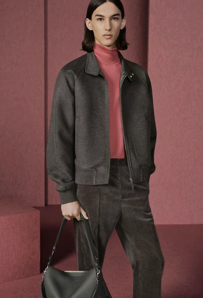 Model wearing a grey zipped jacket and pink turtleneck holding a leather shoulder bag against a textured rose set.