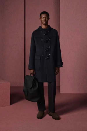 A Black male model stands holding a black leather backpack, wearing a dark hooded duffle coat against a dusty pink studio set.