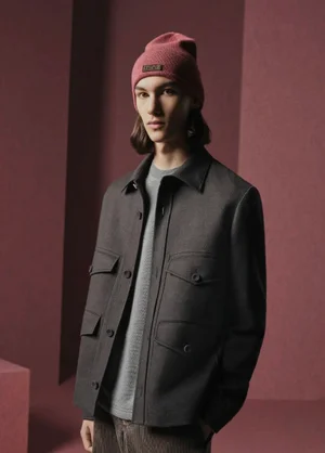 Model wearing a pink beanie and dark buttoned jacket standing against a muted red set-dressed background for Dior Men.