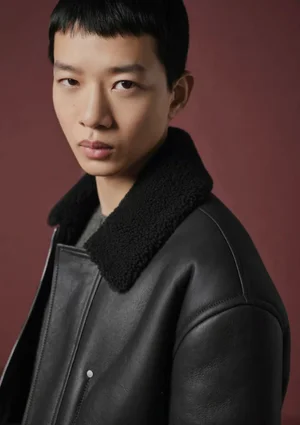 Model Guo Jike wearing a black leather jacket with a plush shearling collar against a deep burgundy background for Dior Men.