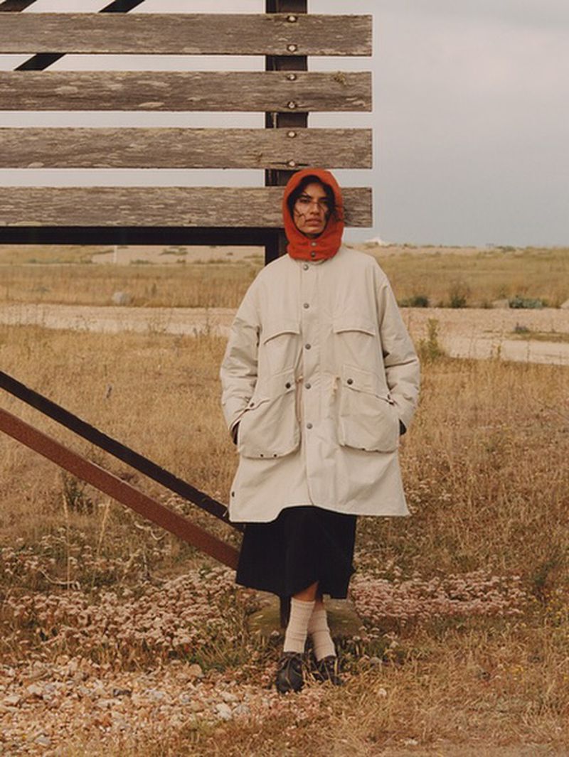 Adult woman stands in a field next to a wooden fence wearing a beige overcoat and orange headscarf. She poses in a rural environment.
