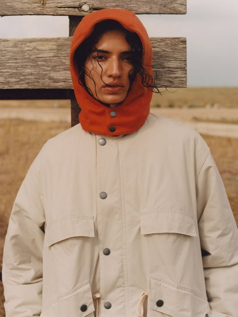 Close-up of a person wearing a beige hooded jacket and red cowl-neck hood with a wooden fence and grassy field backdrop.