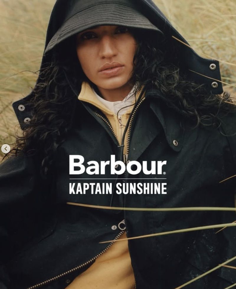 Woman wearing black jacket and bucket hat stands outdoors in a field for a Barbour and Kaptain Sunshine fashion ad.