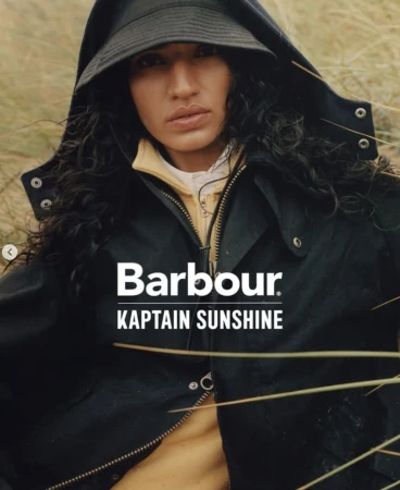 Model wearing a black hooded jacket and bucket hat surrounded by tall dried grass in a Barbour x Kaptain Sunshine campaign.