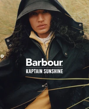 Model wearing a black hooded jacket and bucket hat surrounded by tall dried grass in a Barbour x Kaptain Sunshine campaign.