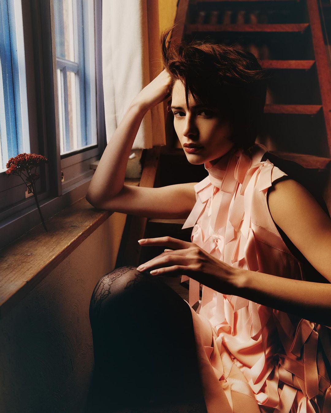 Indoor portrait of a woman in a pink dress with ruffled accents, seated by a window with natural lighting and a ladder in the background.