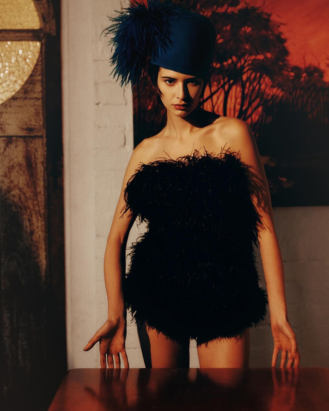 Fashion shot of a woman in a black feathered outfit and blue feathered hat standing indoors in front of an art piece on the wall.