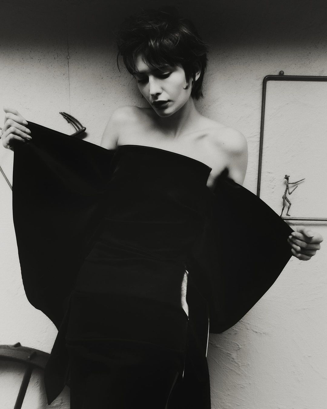 Woman in a strapless black garment holding out fabric edges against a textured wall with framed artwork.