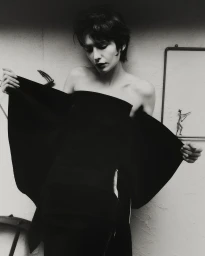 Woman in a strapless black garment holding out fabric edges against a textured wall with framed artwork.