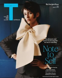 A fashion magazine cover featuring a model wearing a dark blazer with a large cream bow collar against a dark blue paneled wall.