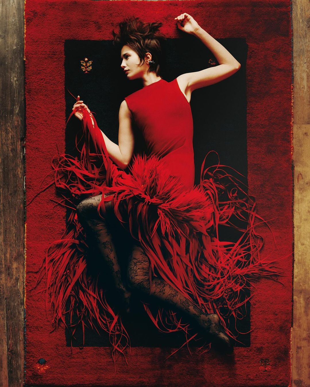 Woman in a red sleeveless dress and black lace tights lying on a textured red rug for T Magazine Style Issue