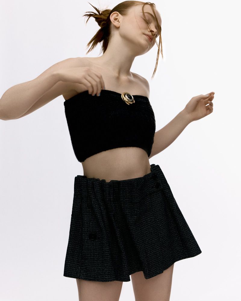 Young woman in a black crop top and pleated mini skirt poses against a white background in a fashion or beauty editorial style.
