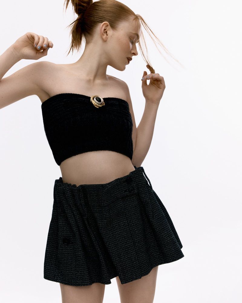 A woman with red hair poses in a black tube top and mini skirt against a white backdrop in a fashion photography studio setting.