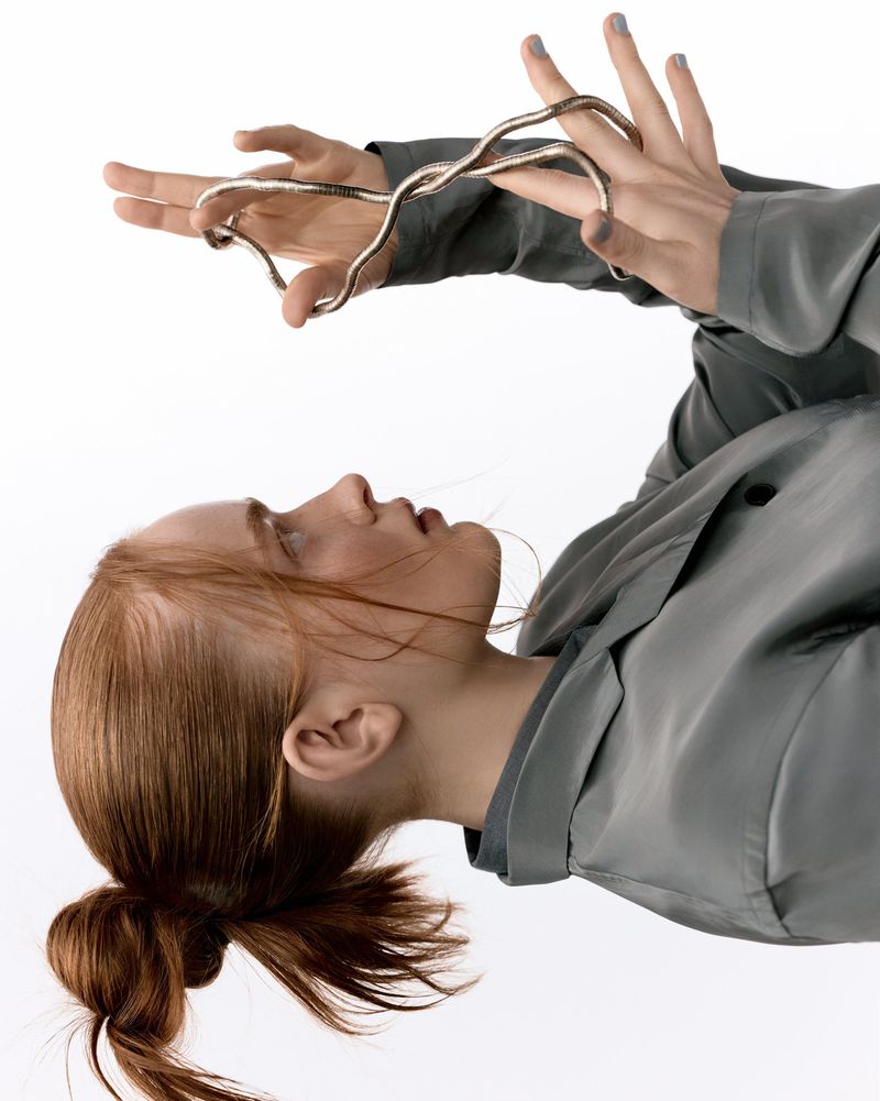 Woman with red hair holds silver, abstract jewelry in a studio shot against a white background, showcasing a clean and minimalist aesthetic.