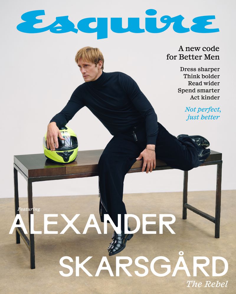 Alexander Skarsgård poses on a table with a helmet for Esquire magazine cover. He wears a black turtleneck and trousers with a light background.