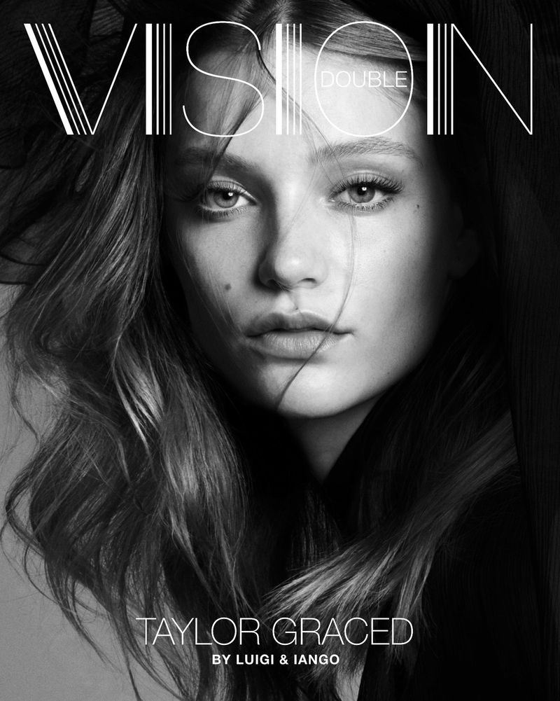 Black and white fashion portrait of a young female model with long wavy hair looking directly at the camera, wearing dark clothing.