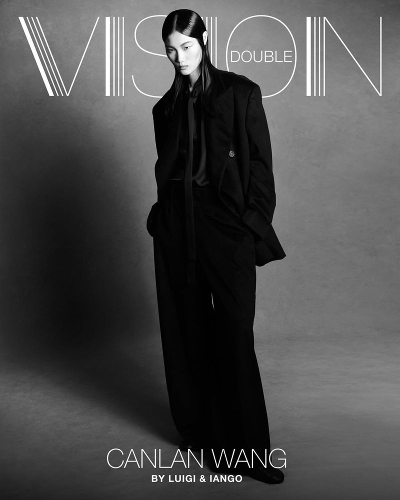 Black and white studio portrait of an Asian fashion model in a dark suit with wide-legged trousers against a gradient backdrop.