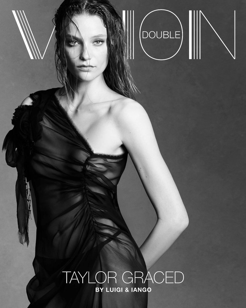 Black-and-white fashion-magazine-style shot of a fair-skinned woman in a semi-sheer black dress, photographed indoors against a neutral background.