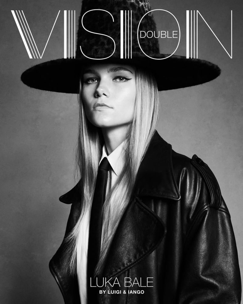 Black-and-white fashion portrait of a woman with long blonde hair, wearing a hat, tie, and trench coat, against a grey backdrop.