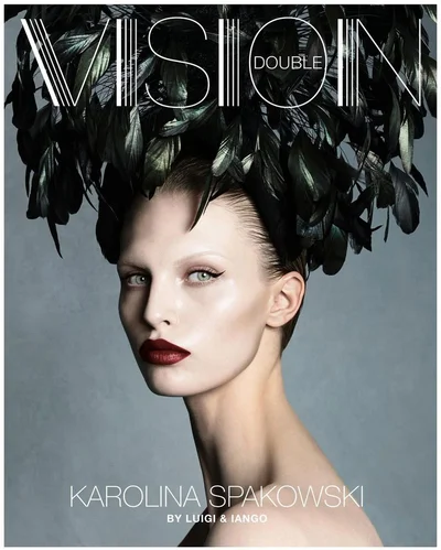 Model Karolina Spakowski wearing an elaborate black feather headpiece and deep burgundy lipstick against a grey backdrop.