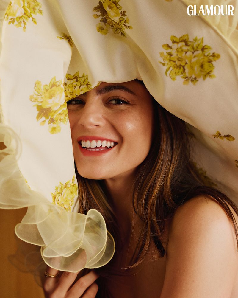 Close-up portrait of Monica Barbaro smiling beneath a large yellow floral fabric brim for Glamour Spain March 2025 covers.
