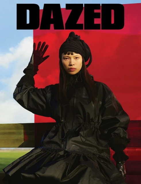 Fashion editorial shot of an Asian woman in a black outfit with a headscarf, posing in front of a red and blue background, with the word 'DAZED' above.