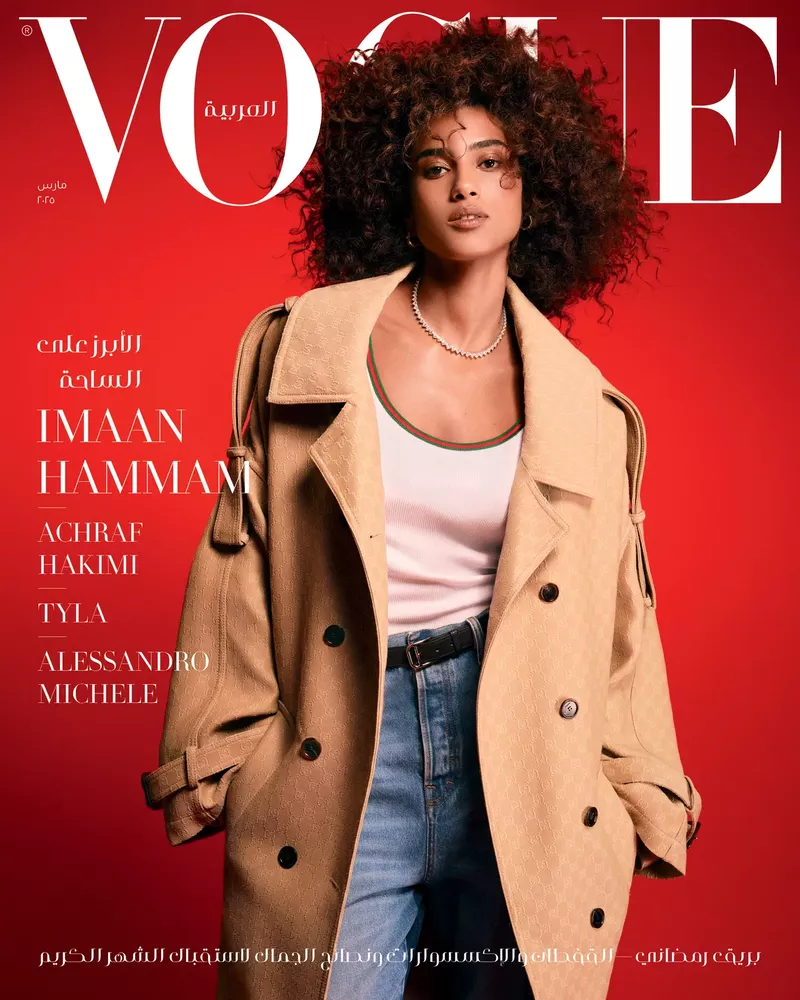 Imaan Hammam stars on Vogue Arabia cover in a tan trench coat, white top, jeans, and a delicate necklace against a red background.