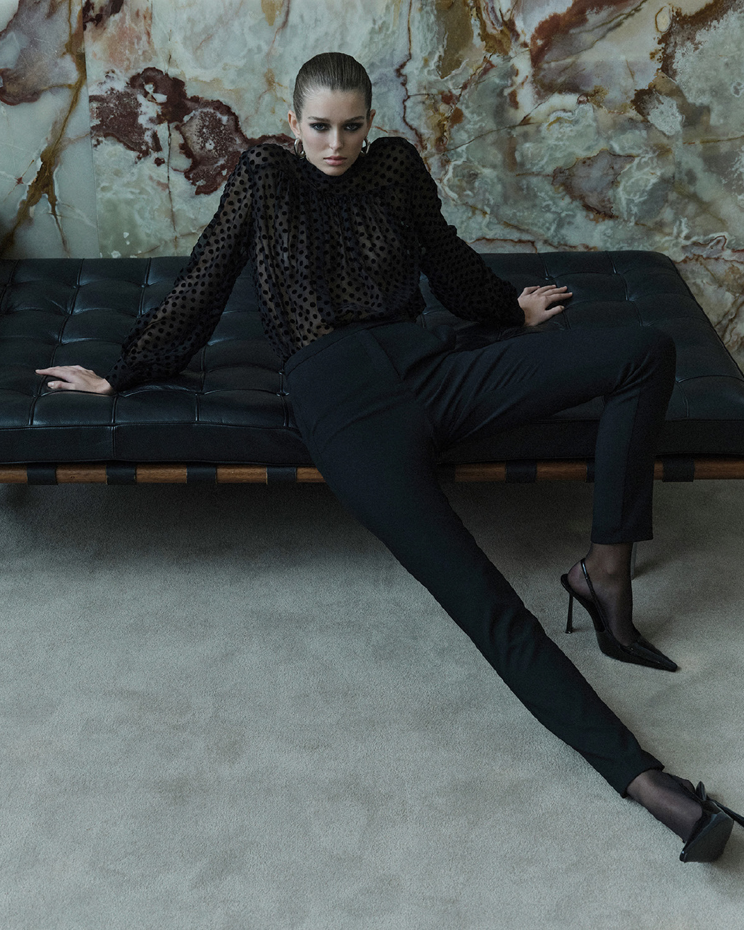 Elegant adult female fashion model poses in a sheer black polka dot top and black pants on a black couch in an indoor setting.