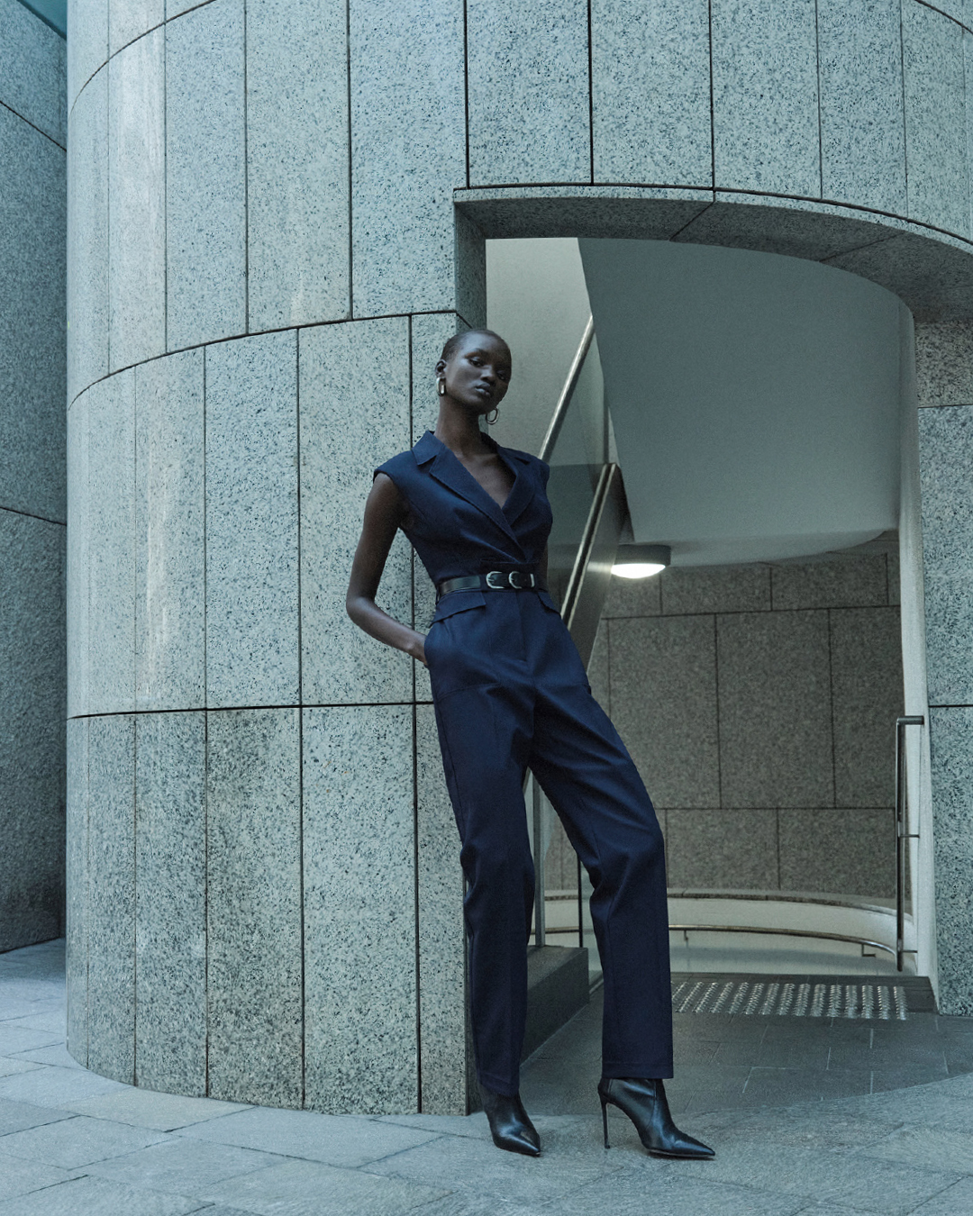 Fashionable Black woman in a dark blue sleeveless jumpsuit and high heel boots poses in an architectural setting with geometric features.