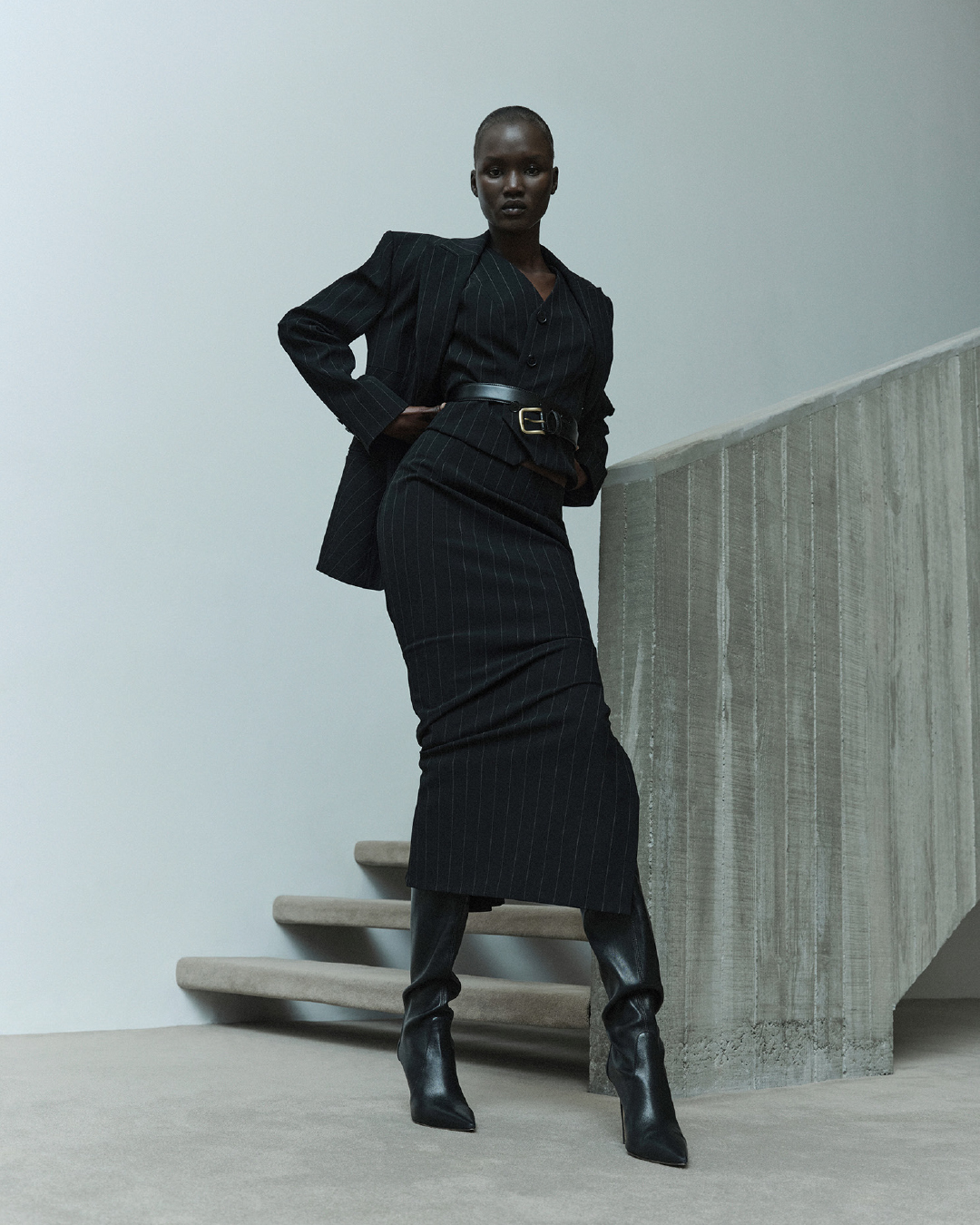 Elegant Black Woman in Pinstripe Suit, Vest, Skirt and Boots Posing Near Concrete Wall Inside Fashion Photo Studio