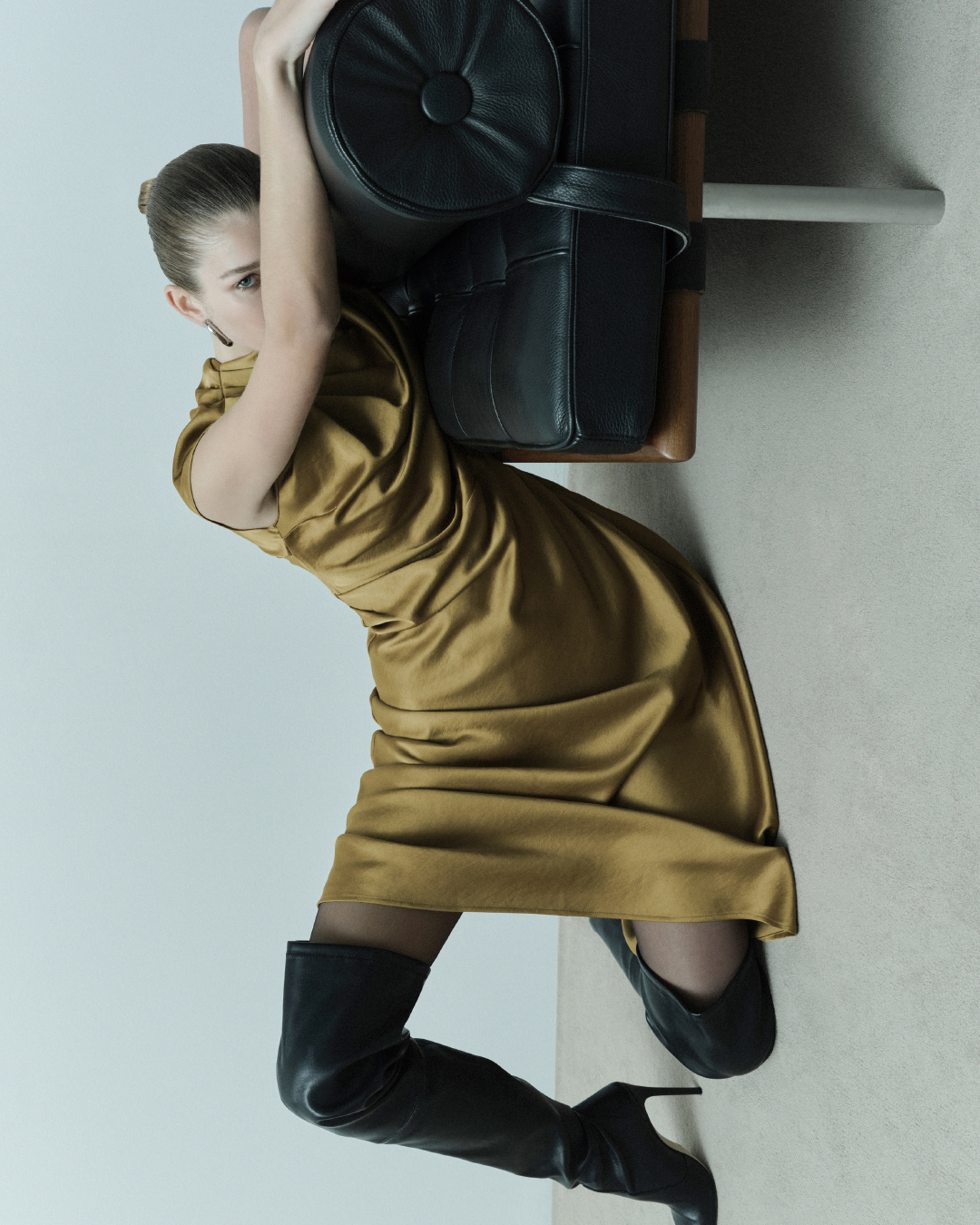 Woman in gold dress and black boots poses with modern black leather chair in a minimalist studio setting. Fashion editorial shot.