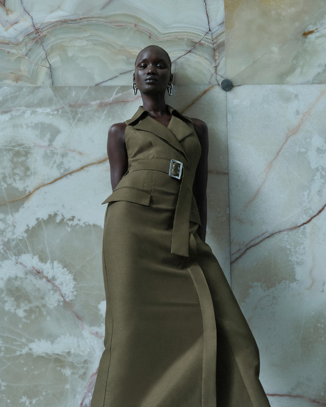 A bald Black woman wearing an olive green sleeveless belted dress posing against a patterned marble wall.