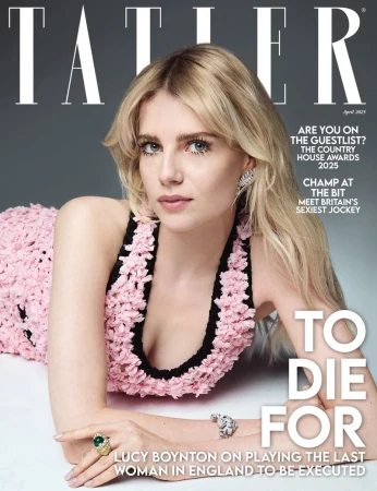 Lucy Boynton reclining wearing a pink textured dress with black trim against a grey studio background for a magazine cover