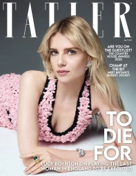 Lucy Boynton reclining wearing a pink textured dress with black trim against a grey studio background for a magazine cover
