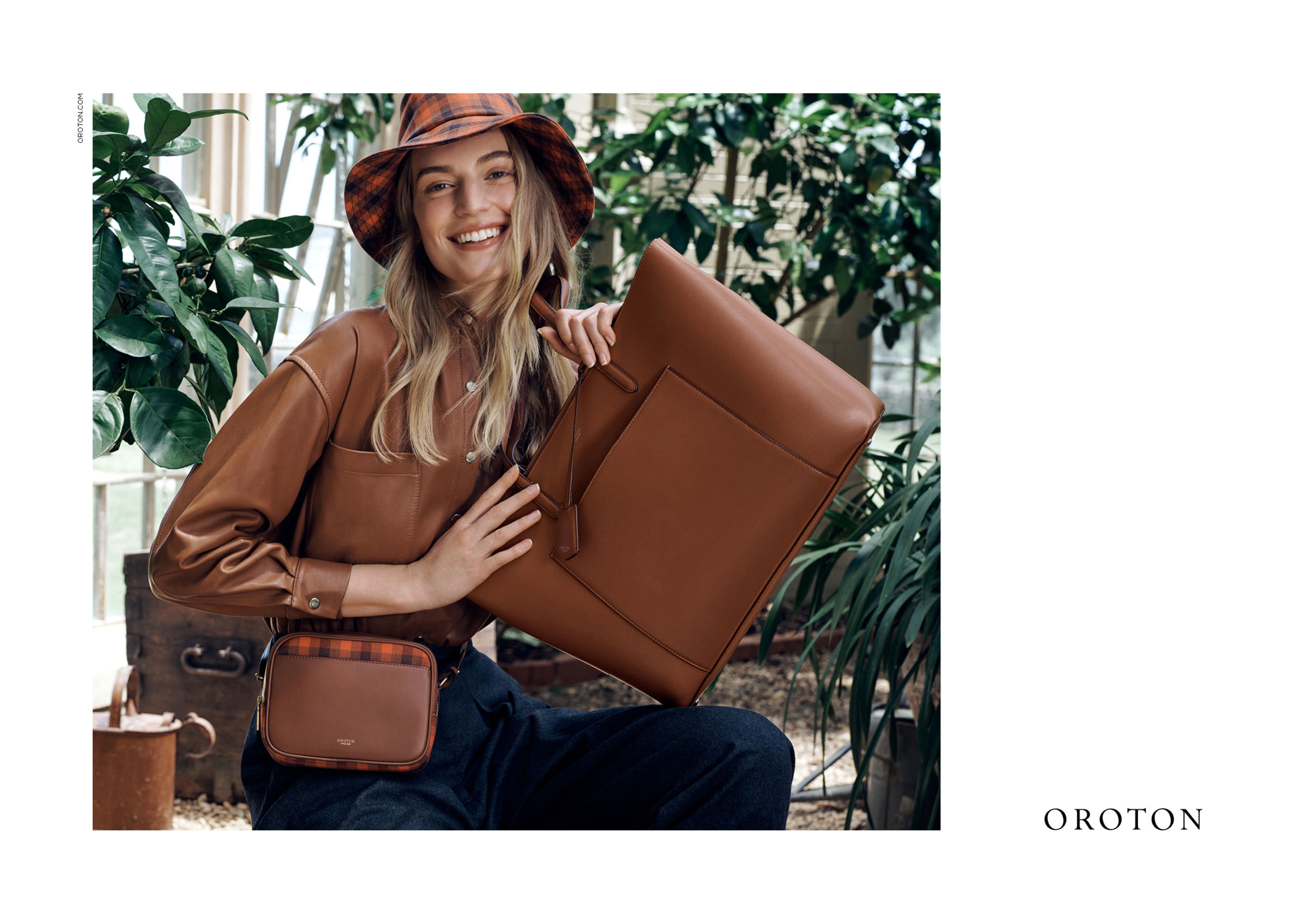 Smiling adult female model poses with a brown leather tote bag and purse in a bright indoor setting with green foliage visible.